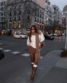Woman in a brown skirt, white sweater, and tall boots on a city street