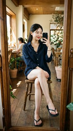 Woman in a dark collared shirt and flip flops taking a mirror selfie indoors