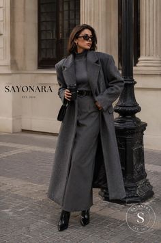 Woman in a long gray coat and turtleneck holding a coffee cup on a street