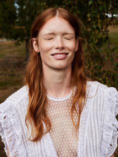 Woman with red hair and freckles smiling with eyes closed in a white lace top