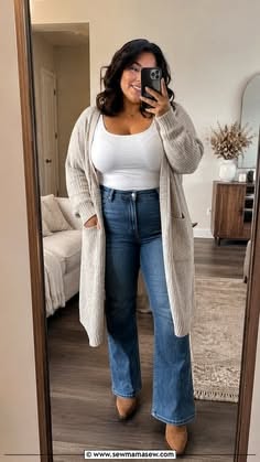 Woman in a long cardigan, white top, and blue jeans taking a mirror selfie