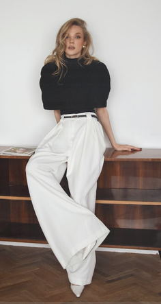 Woman with blonde hair wearing a black top and white high-waisted pants indoors