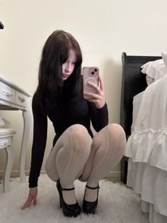 Woman in a black top and white tights taking a mirror selfie in a bedroom