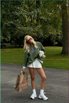 Blonde woman in a green sweatshirt and white skirt holding a shopping bag outdoors
