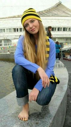 Woman with long blonde hair wearing a yellow beanie and blue shirt, crouching barefoot outdoors