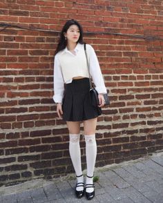 Woman in a white shirt, sweater vest, and striped skirt standing by a brick wall