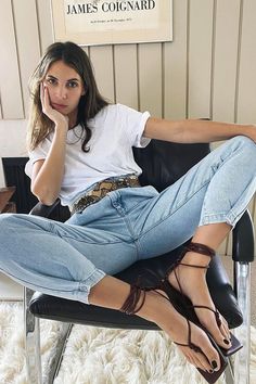 Woman in a white t-shirt and light wash jeans with lace-up sandals sitting indoors
