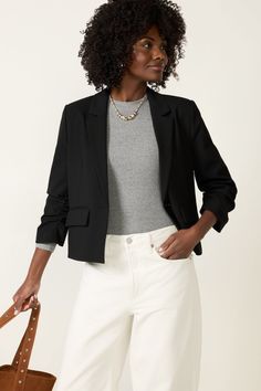 Woman with curly hair wearing a black blazer and white jeans, holding a brown tote bag