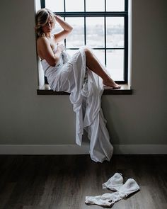 Woman wrapped in a white sheet sitting on a window sill with lingerie on the floor
