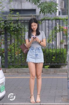 Woman in a denim skirt and grey shirt standing on a street looking at her phone