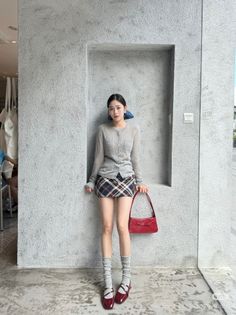 Woman in a plaid skirt and cardigan holding a red handbag indoors