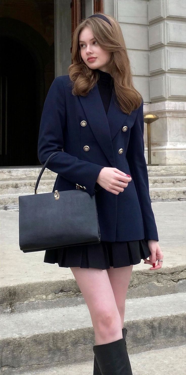 Woman in a navy blazer and black pleated skirt holding a handbag outdoors