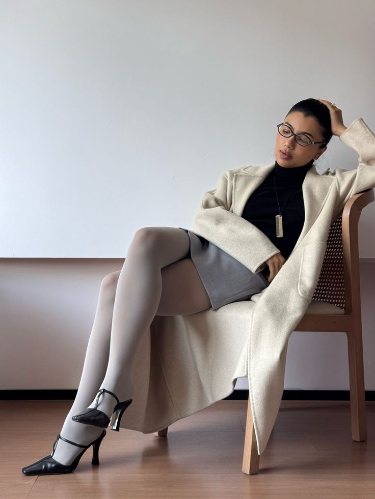 Woman in a long coat, turtleneck, mini skirt, and white tights sitting on a chair
