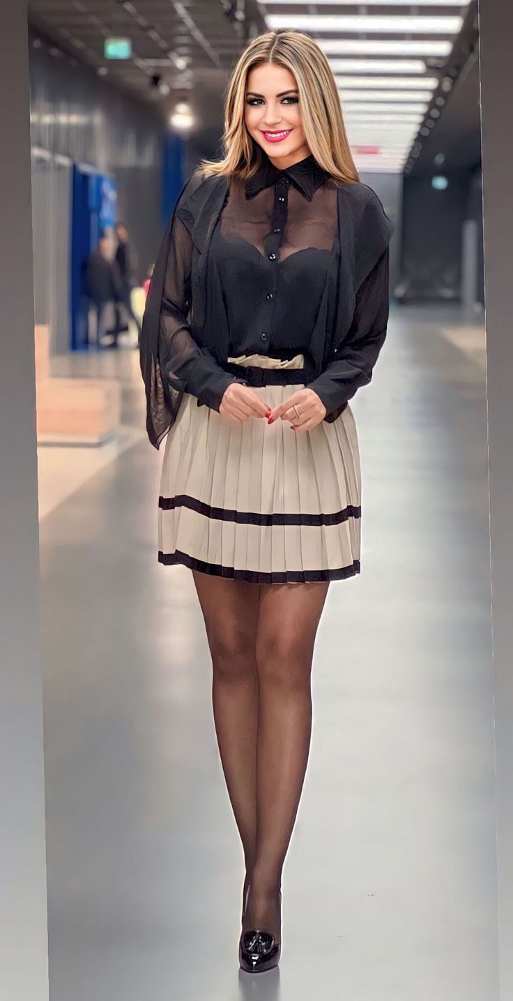 Woman in a sheer black blouse and pleated skirt smiling indoors