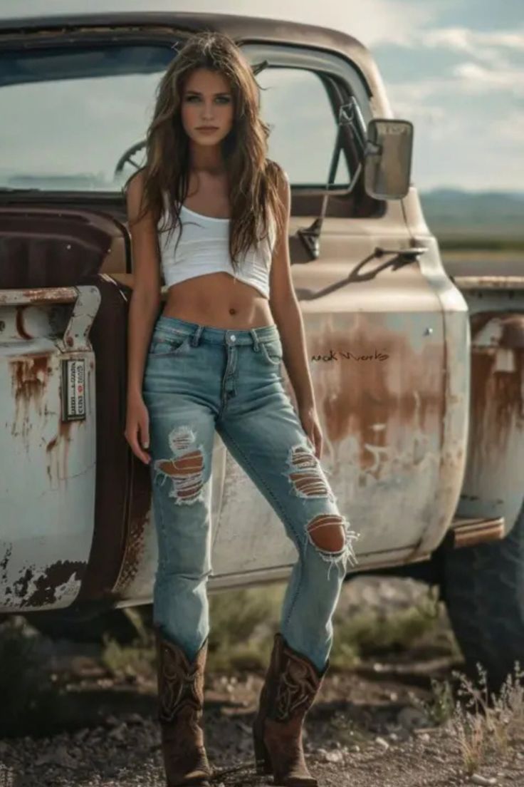 Woman in a crop top, ripped jeans, and cowboy boots leaning on an old truck
