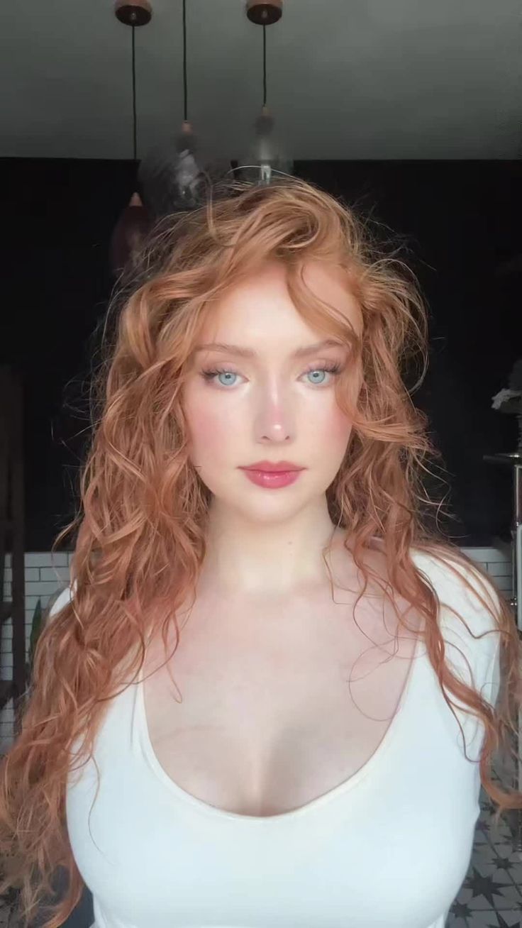 Woman with long curly red hair and blue eyes wearing a white top indoors