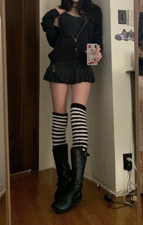 Person in a black hoodie, pleated skirt, and striped knee-high socks taking a mirror selfie
