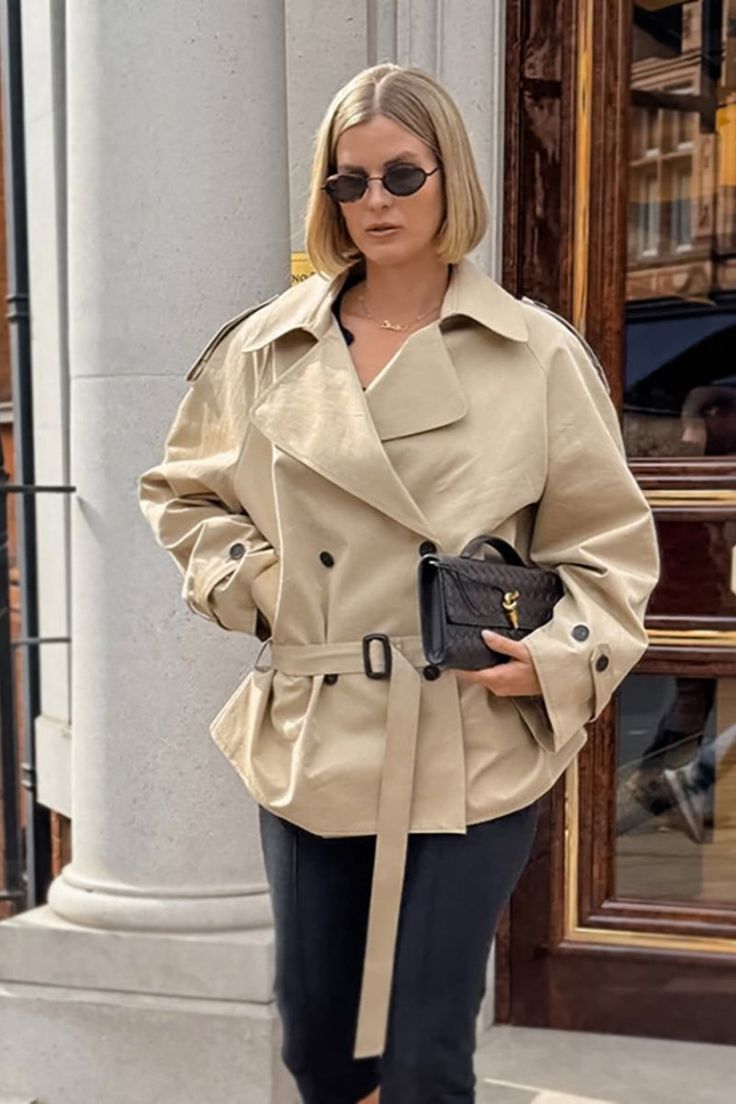Woman in a beige trench coat and sunglasses holding a black handbag outdoors