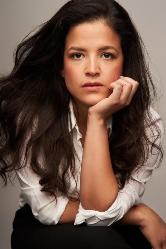 Woman with long dark hair wearing a white shirt, resting her chin on her hand