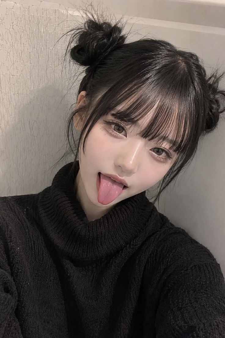 Woman with space buns and bangs sticking out her tongue in a black sweater