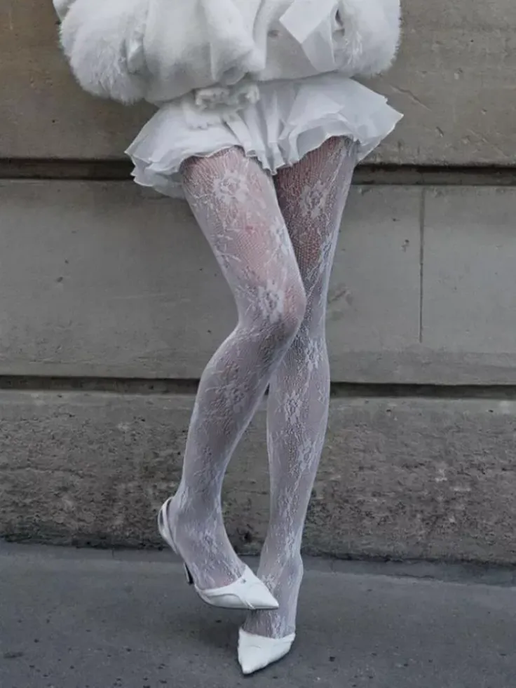 Person wearing white lace tights, a white skirt, and white heels against a concrete wall