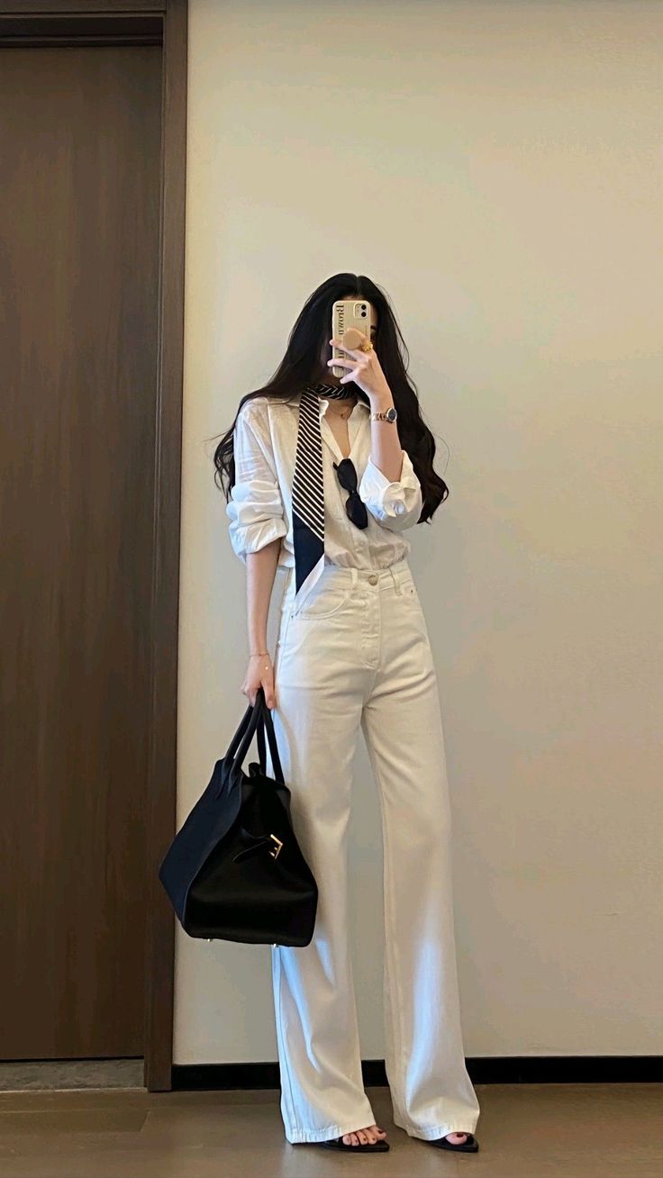 Woman in a white shirt and pants holding a black handbag, taking a mirror selfie
