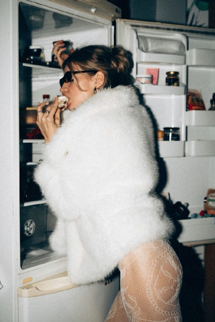 Woman in a white fur coat and lace tights eating from a refrigerator