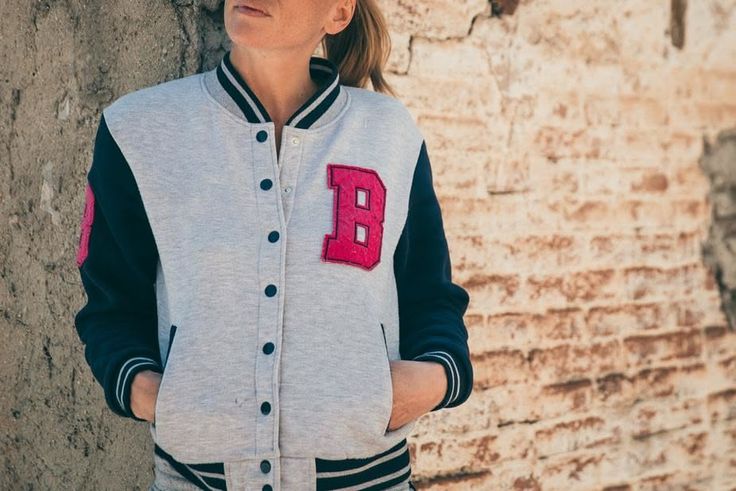 Woman in a varsity jacket with hands in pockets, leaning against a brick wall