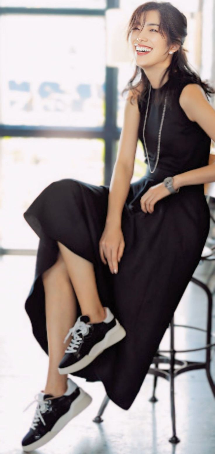 Woman in a black dress and sneakers, smiling while sitting indoors