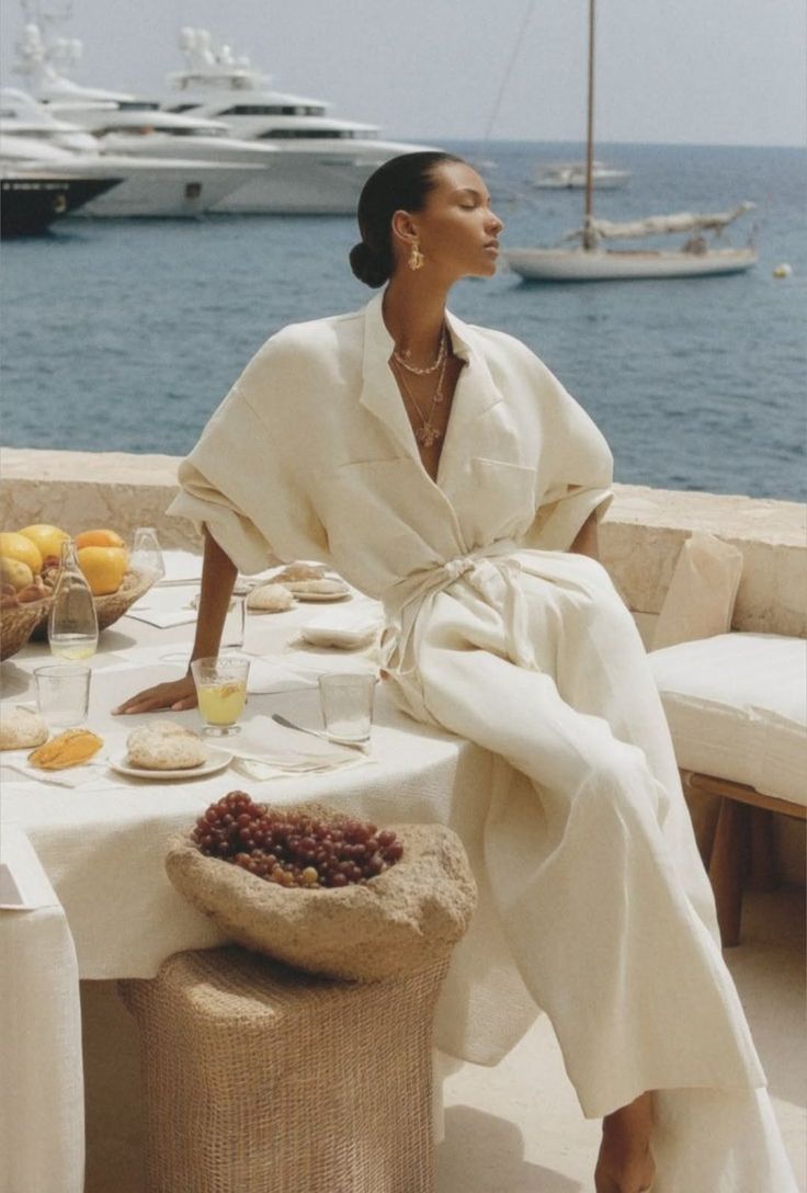 Woman in a white jumpsuit dining outdoors with an ocean view and yachts