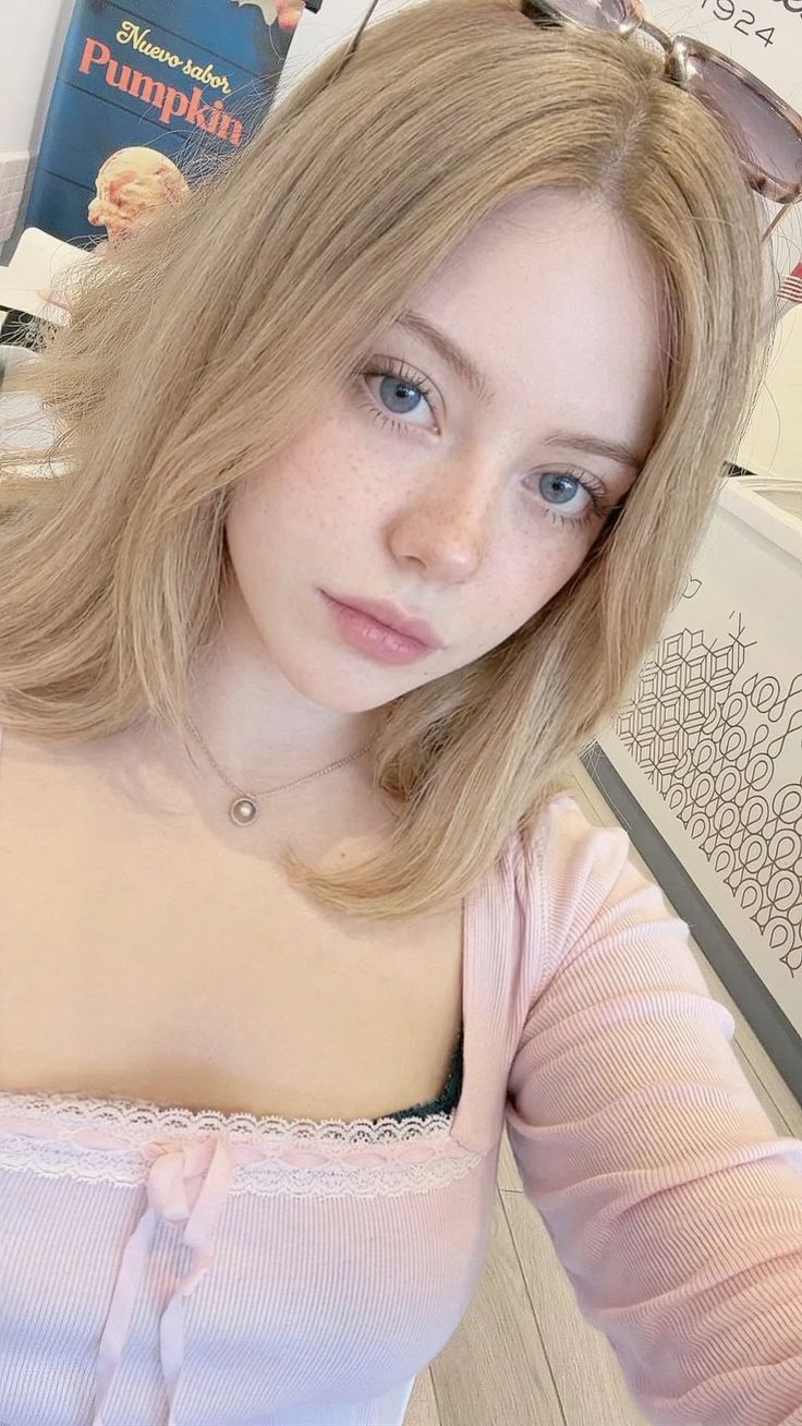 Blonde woman with blue eyes and freckles wearing a pink top with lace trim