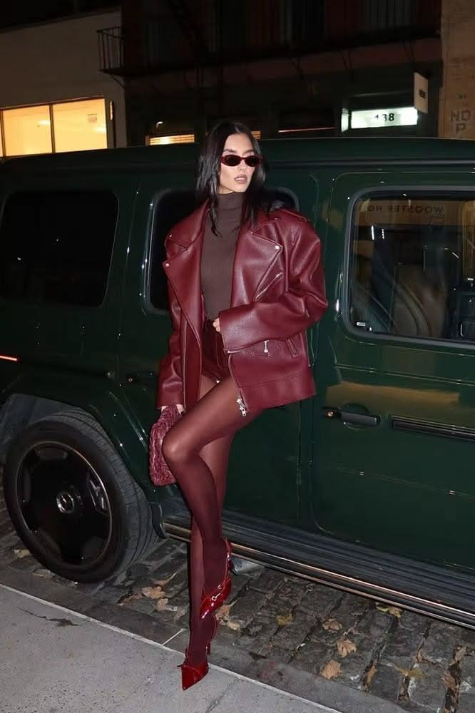 Woman in a red leather jacket and sunglasses posing next to a dark green car at night