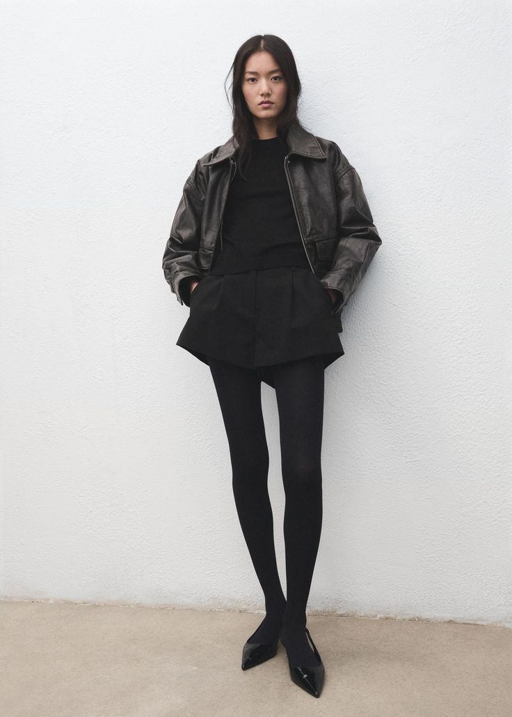 Woman in a black leather jacket, shorts, and tights against a white wall