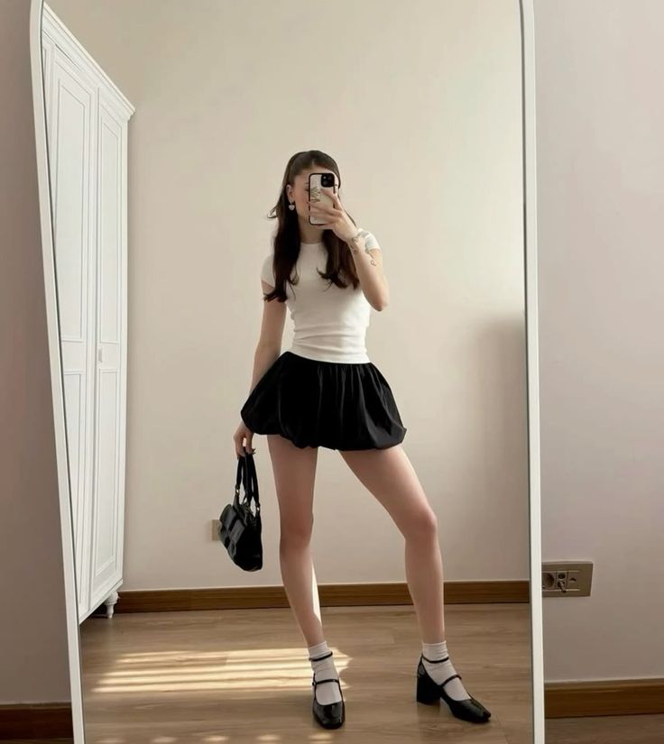 Woman in a black bubble skirt and white t-shirt taking a mirror selfie indoors