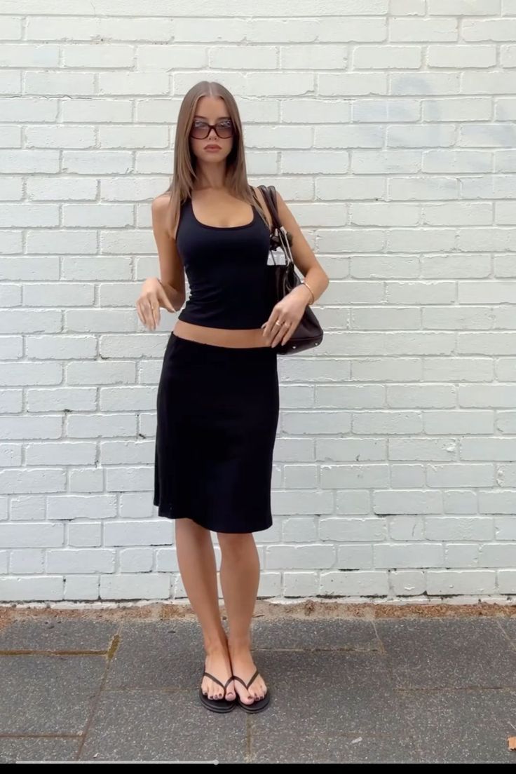 Woman in a black tank top and midi skirt posing against a white brick wall