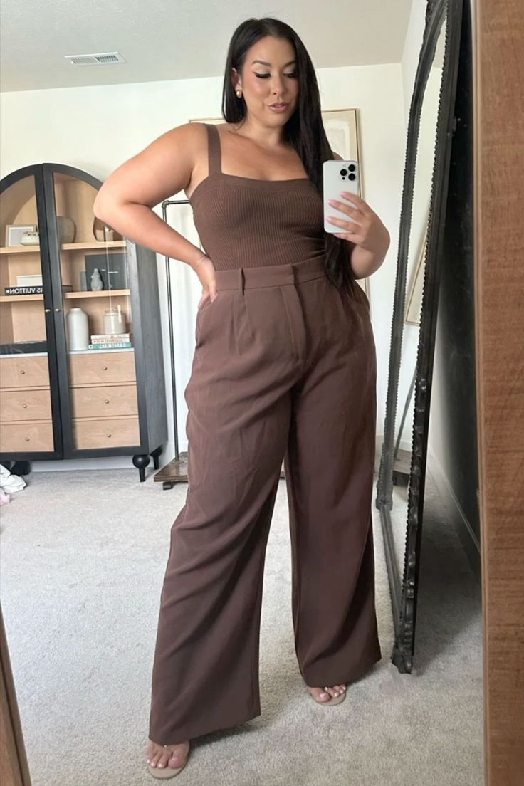 Woman with long dark hair in a brown top and wide leg pants taking a mirror selfie
