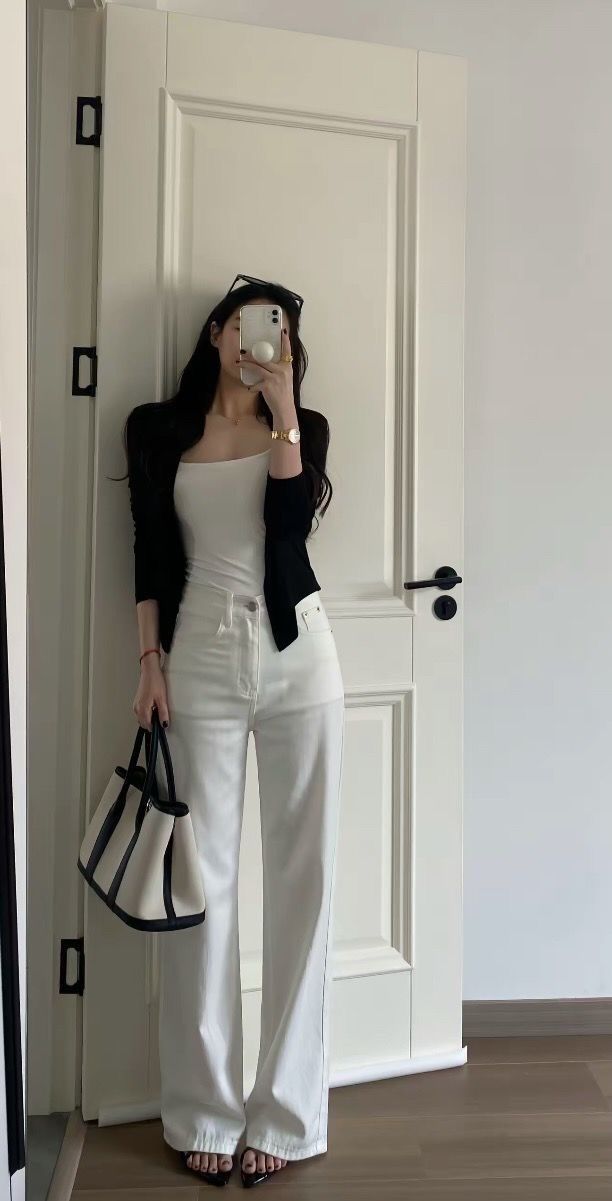 Woman in a white tank top and pants with a black cardigan taking a mirror selfie