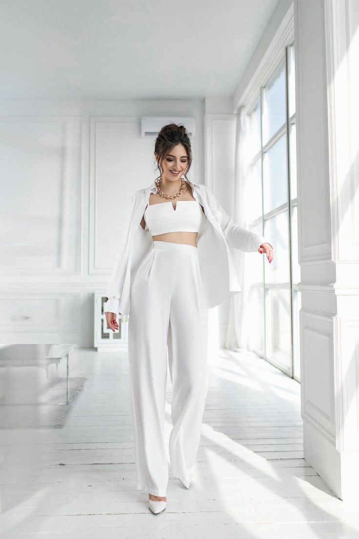 Woman in a white crop top and wide leg pants with a gold necklace indoors
