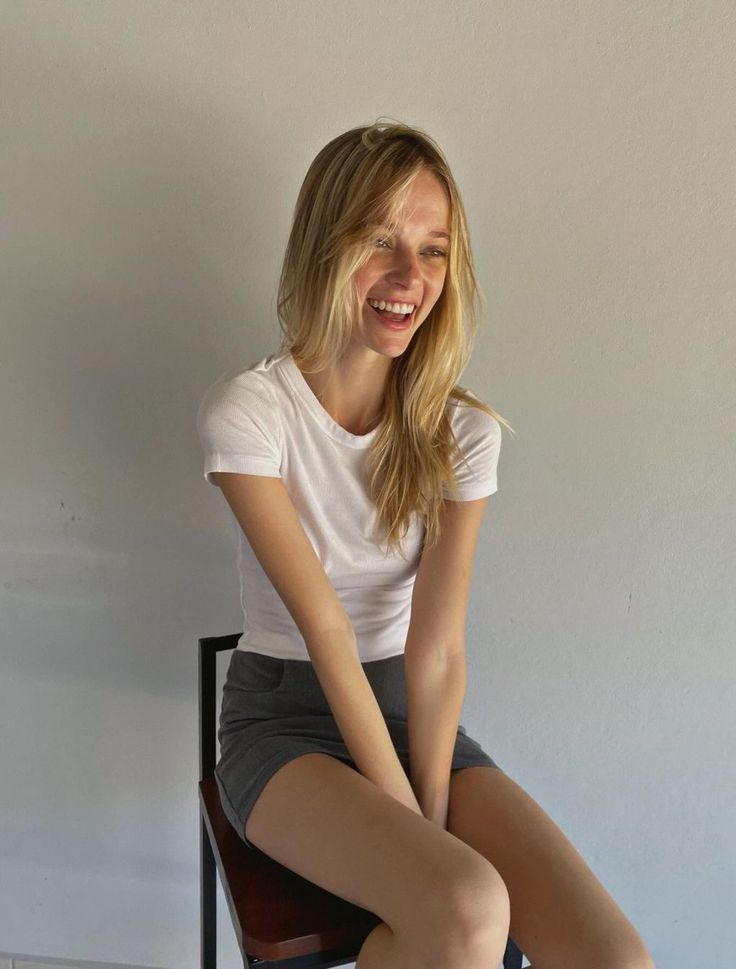 Blonde woman in a white t-shirt and grey skirt sitting indoors and smiling