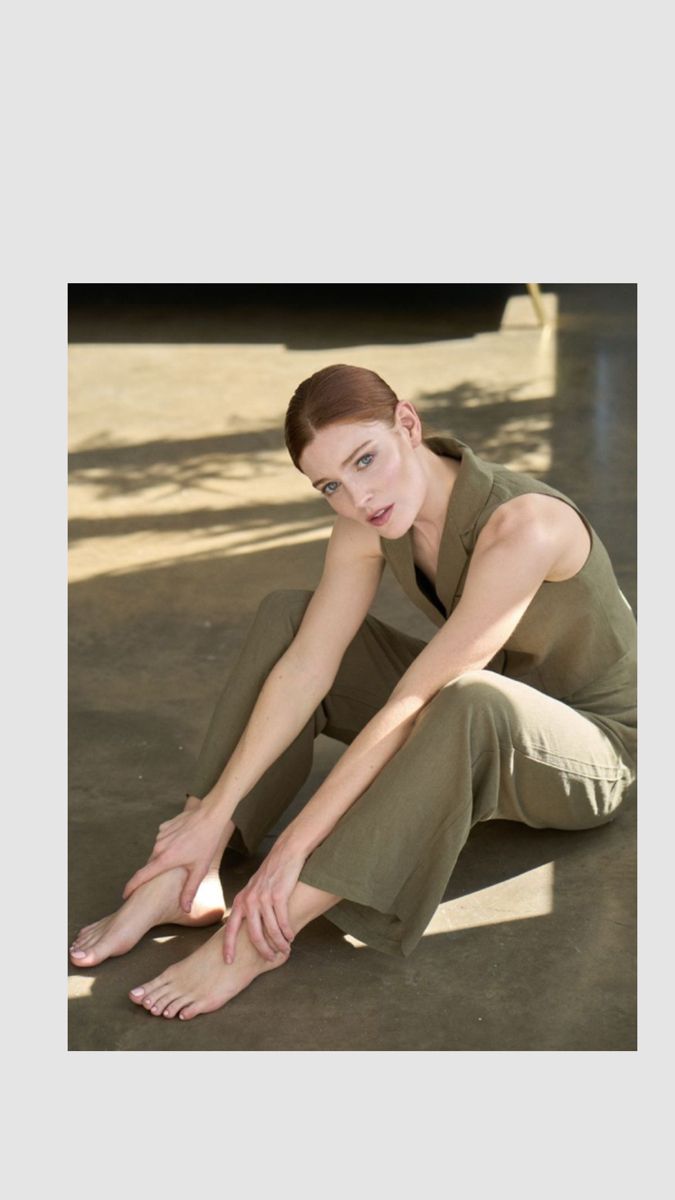 Woman with red hair in a sleeveless top and wide-leg pants sitting barefoot indoors