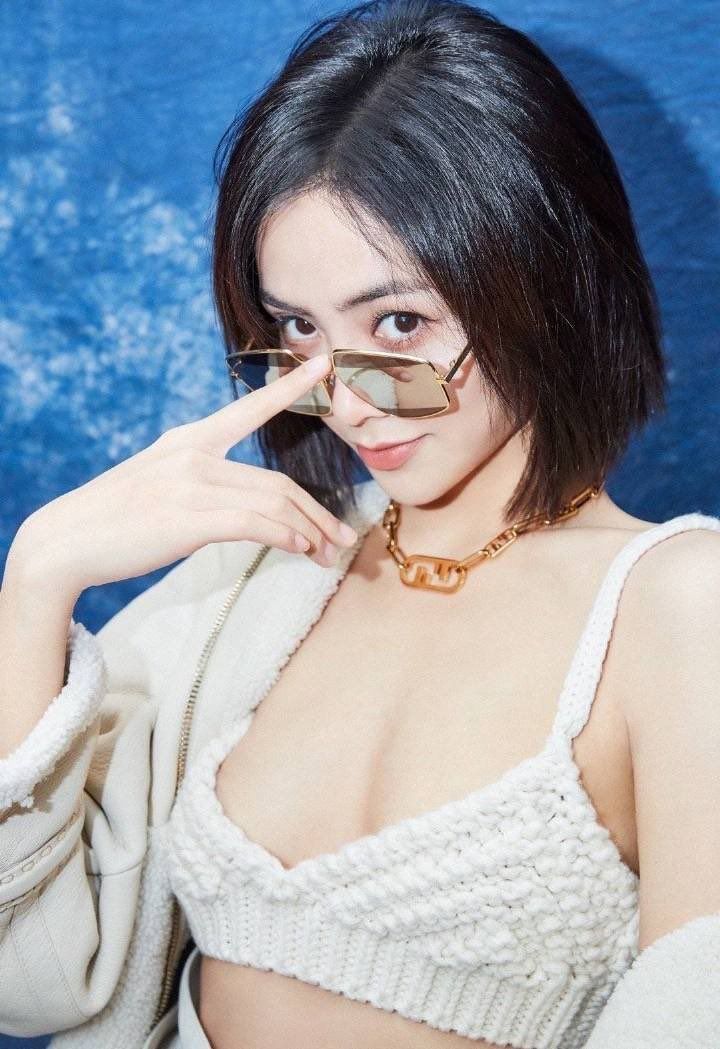 Woman with short hair and sunglasses wearing a white top and choker against a blue background