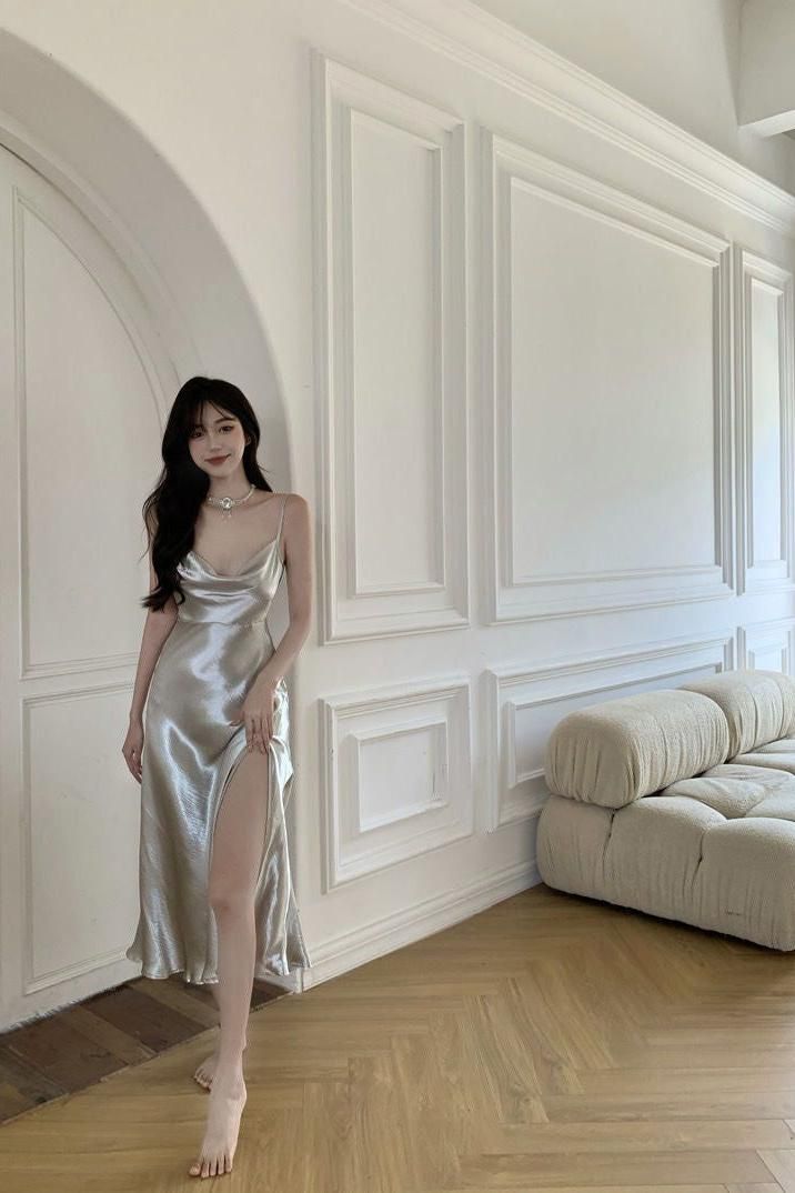 Woman in a silver cowl neck dress with a high slit standing barefoot indoors