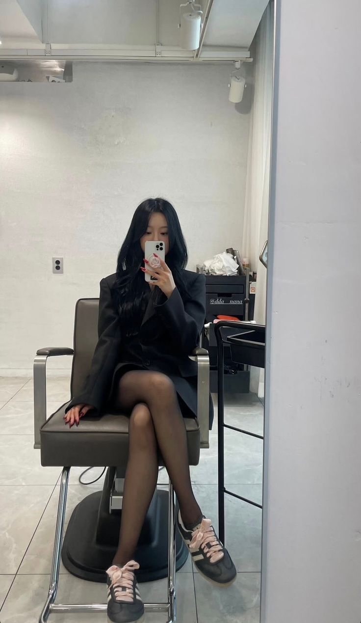 Woman in a black blazer and tights taking a mirror selfie while sitting
