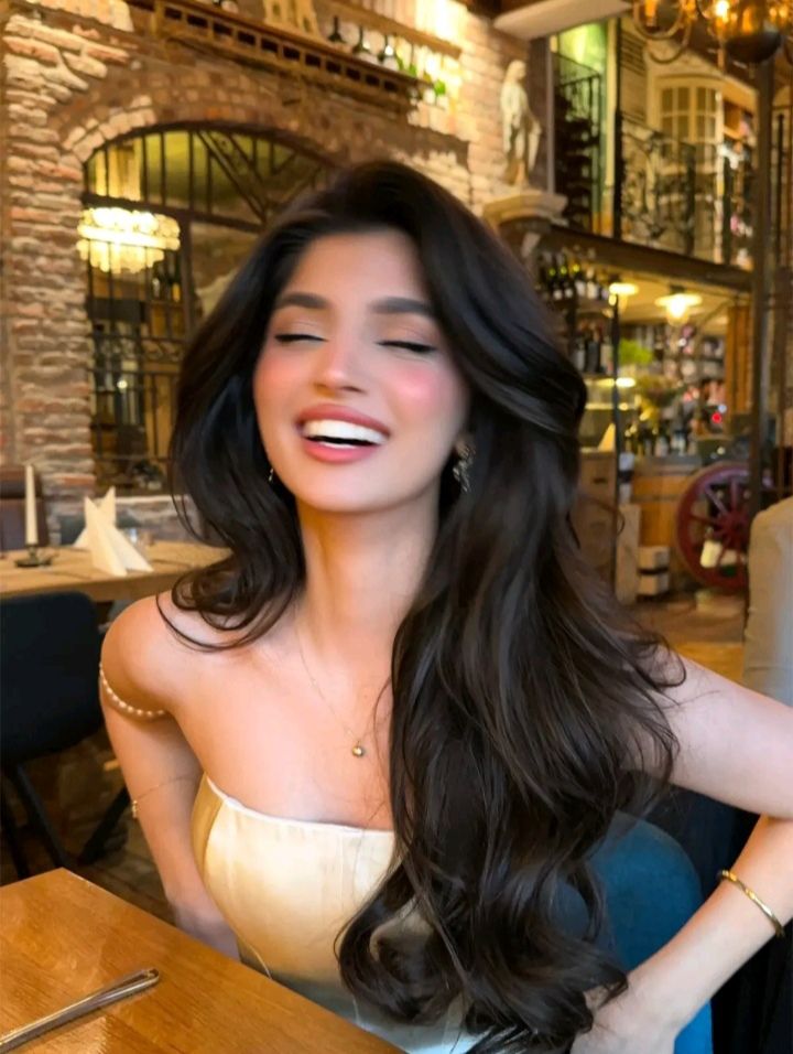Woman with long dark hair wearing a strapless top and pearl necklace, smiling in a restaurant