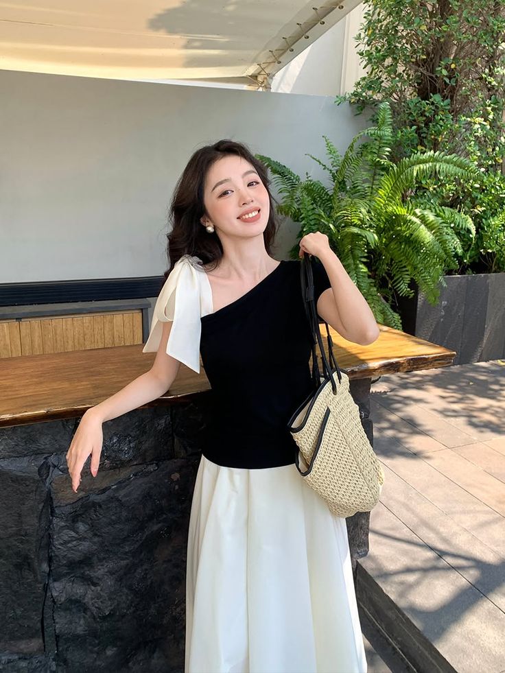 Woman in a black one-shoulder top and white skirt holding a straw bag outdoors
