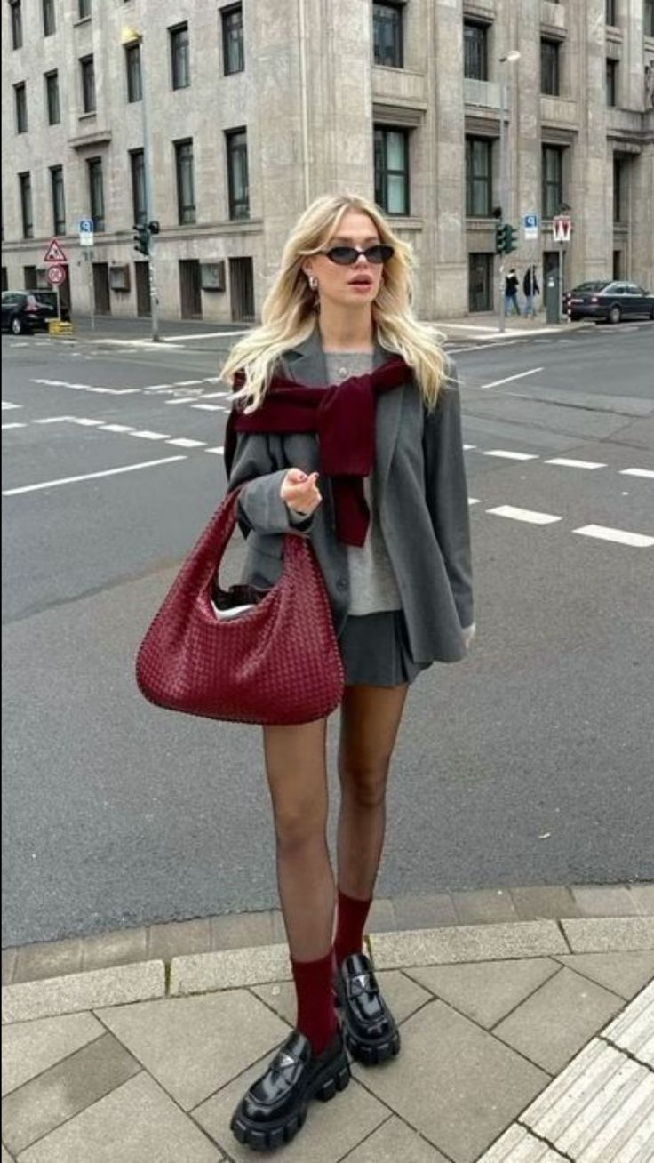 Woman with blonde hair in a gray blazer and mini skirt holding a red bag on a city street