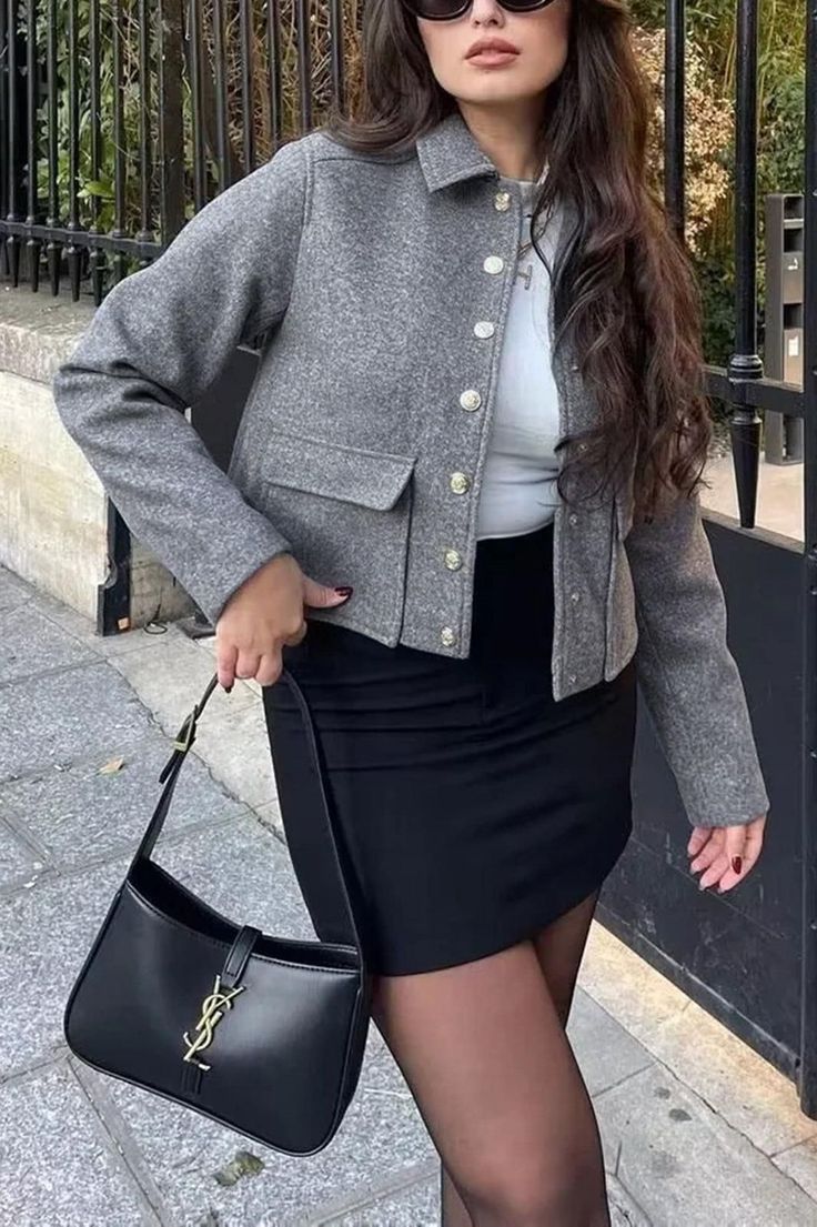 Woman in a gray jacket and black mini skirt holding a black handbag outdoors