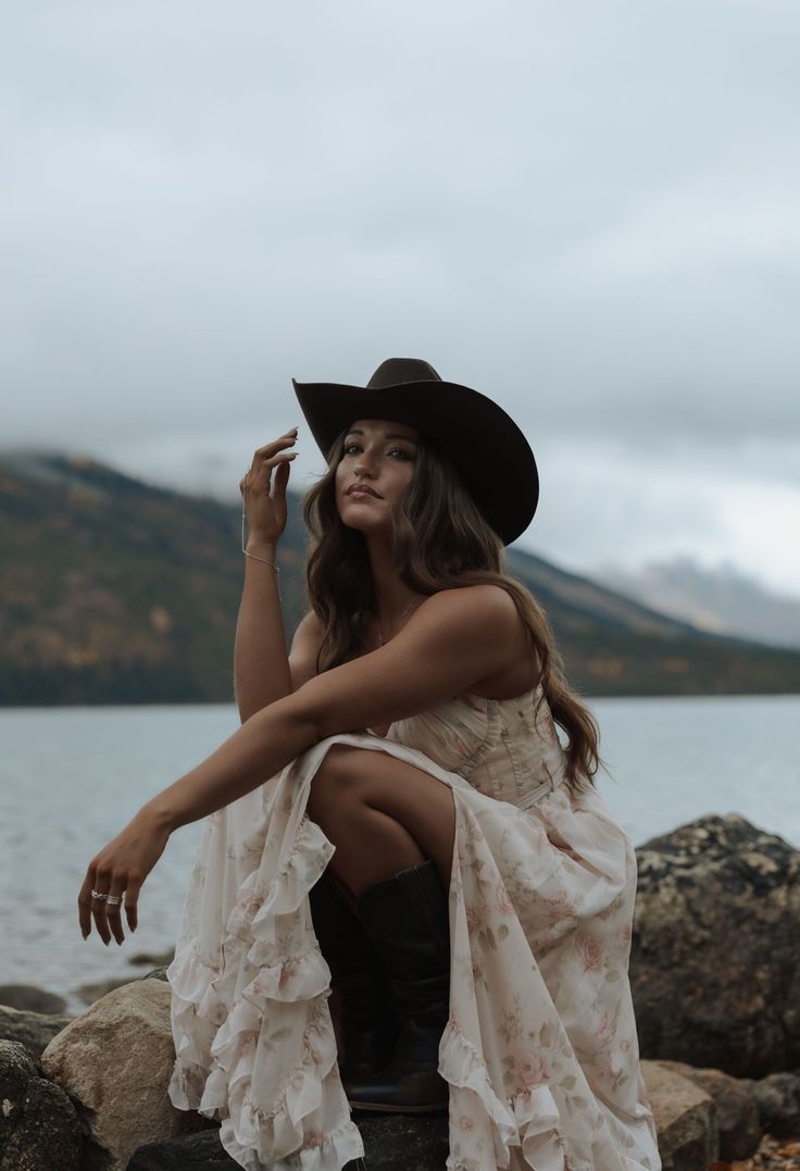Woman in a cowboy hat and floral dress crouching by a lake with mountains in the background