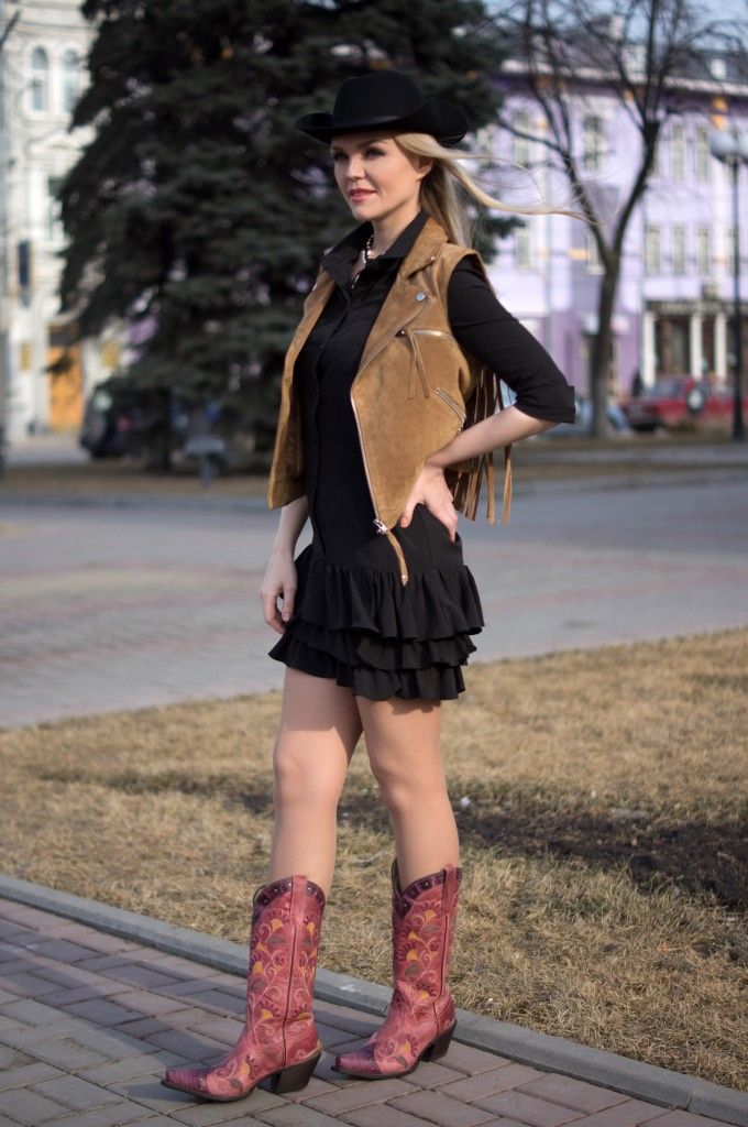 Woman in a cowboy hat, fringe vest, and black dress with cowboy boots outdoors