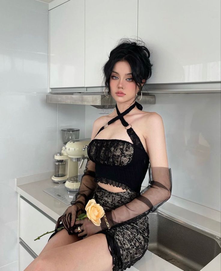 Woman with black hair in a lace outfit and sheer gloves holding a flower in a kitchen